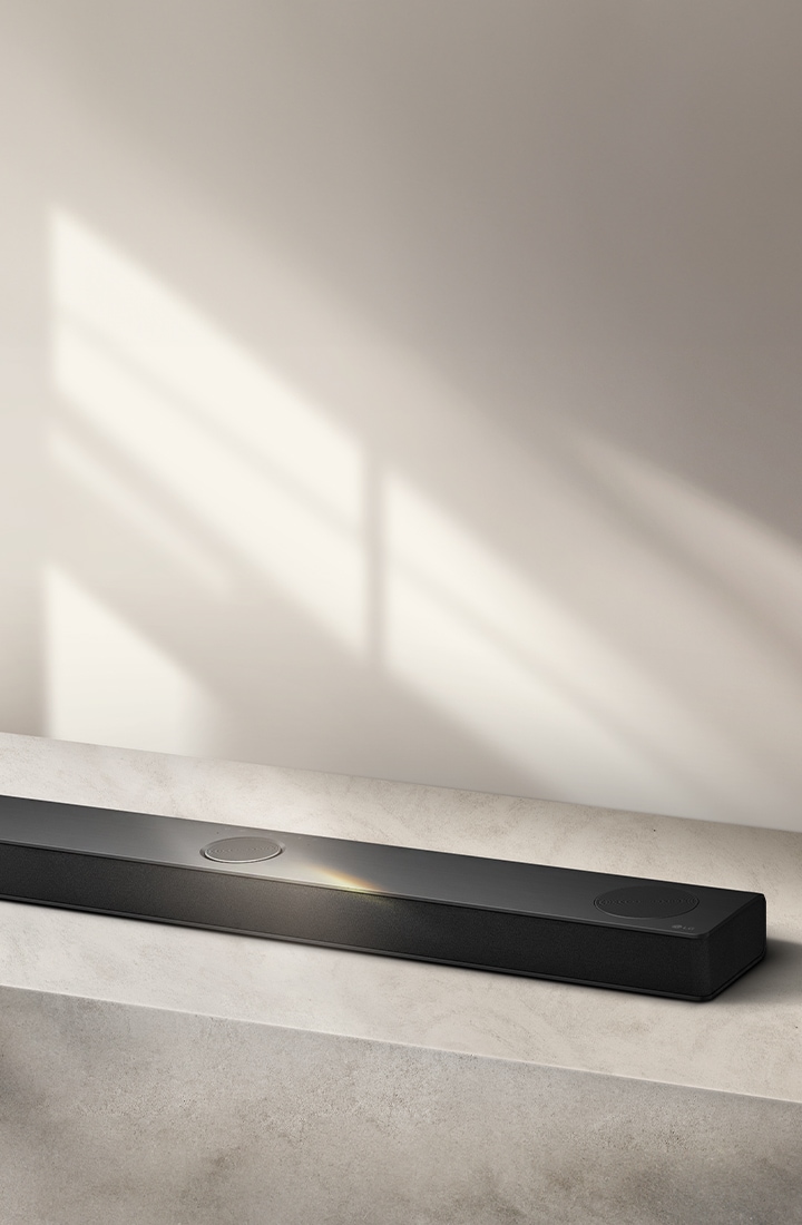 LG Soundbar จัดวางบนพื้นผิวสีเทา ฉากหลังโทนสีเทา มุมมองจากด้านบนแบบเอียง ช่วยขับให้เห็นดีไซน์ที่เรียบและทันสมัย