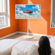 LG StanbyME 2 Max LX6 Movable Screen is placed in a bright room with orange walls beside a window, displaying a red airplane flying across a blue sky, highlighting vivid screen content and flexible placement for relaxed viewing.