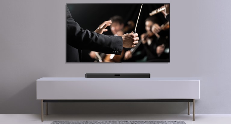 ทีวีปรากฏบนผนังสีเทา และ LG Soundbar อยู่ด้านล่างชั้นวางสีเทา ทีวีแสดงภาพวาทยากรที่กำลังนำวงออเคสตร้า