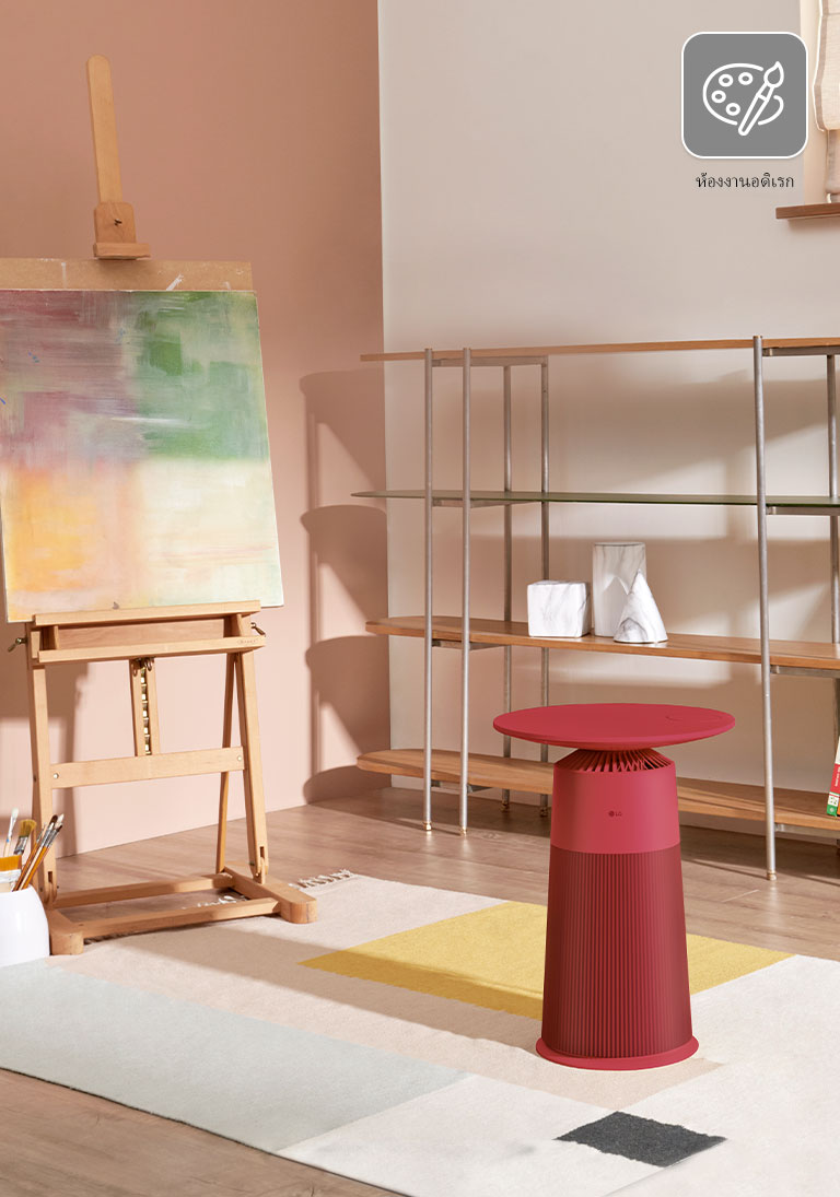 ห้องศิลปะมีขาตั้งวาดภาพด้านซ้าย โต๊ะฟอกอากาศ LG Puricare สีแดงวางไว้กลางห้อง พร้อมชั้นวางหนังสือด้านขวา