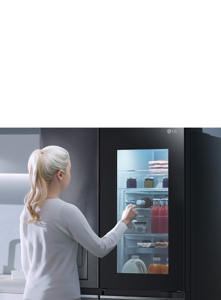 หญิงสาวมองตู้เย็น LG ที่มีประตูโปร่งแสง มองเห็นเครื่องดื่มและของหวานด้านในในครัวสมัยใหม่