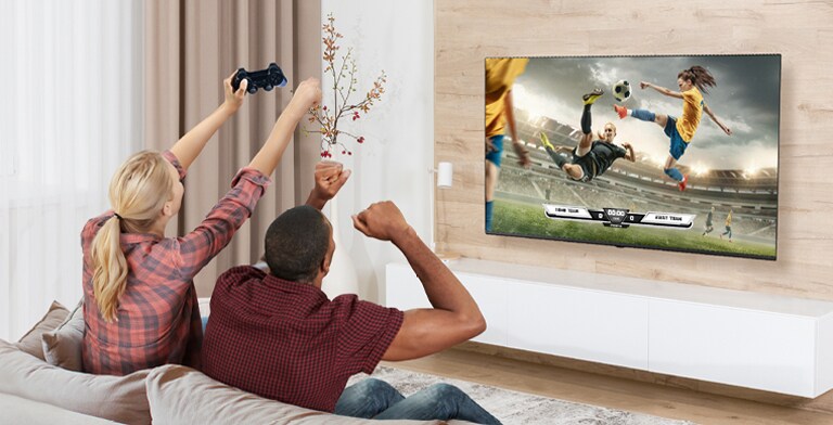 Bu resimde, oyun oynayan bir adam ve kadın yer alıyor ve TV ekranında gerçekçi bir oyun sahnesi gösteriliyor.