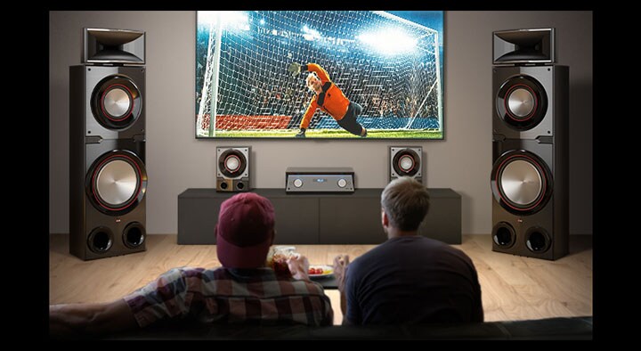 LG MR11 oturma odasında gösteriliyor. İki dev boy hoparlörün ortasında duvara asılı LG TV ekranında bir futbol maçı görüntüleniyor. İki kişi futbol maçını izliyor.