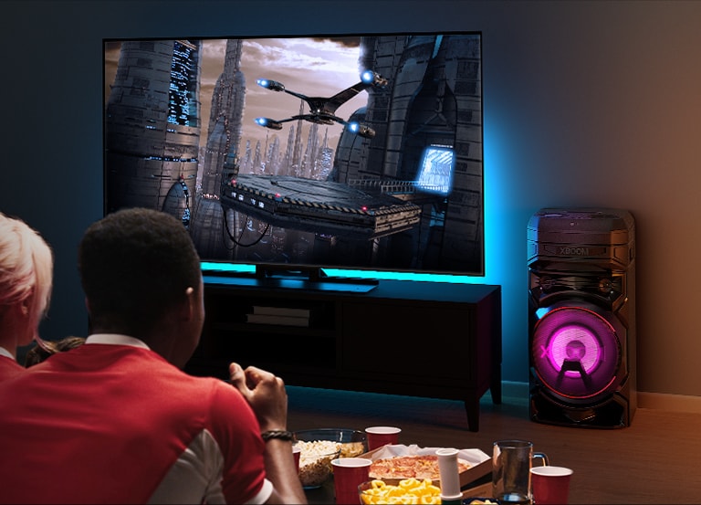People watching TV on a wall with an LG XBOOM RNC5 to the right of it. TV is displaying a movie. 