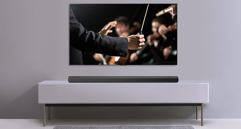Gri bir duvara asılı TV ile altındaki gri rafta duran LG Soundbar. Orkestrayı yöneten orkestra şefini gösteren TV.