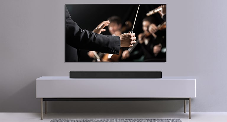 Gri bir duvara asılı TV ile altındaki gri rafta duran LG Soundbar. Orkestrayı yöneten orkestra şefini gösteren TV.