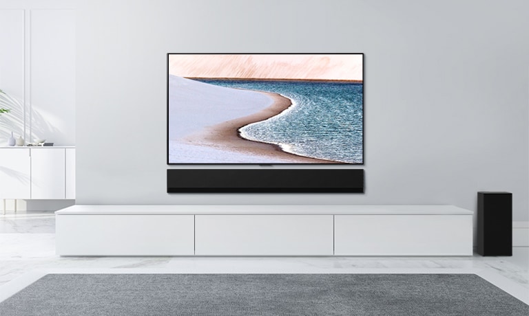 Açık gri duvara monte edilmiş TV. Alttaki beyaz dolabın üzerinde LG Soundbar. Sahil görüntüsü olan TV.