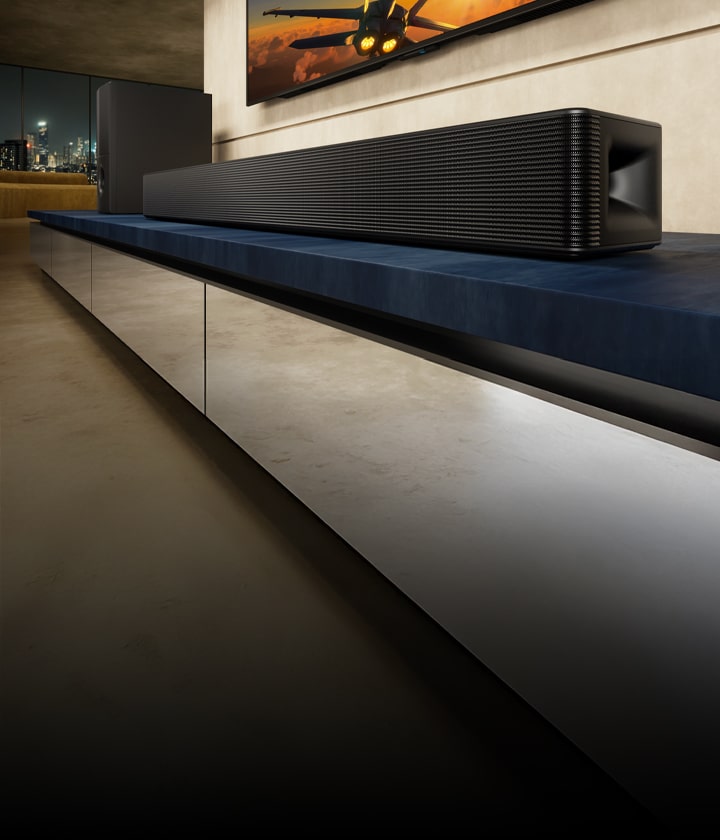 LG Soundbar SH5A ve subwoofer, oturma odasında mavi bir TV ünitesinin üzerinde duruyor. Uçak görüntüsü olan TV, soundbar’ın üzerinde kısmen görünüyor.