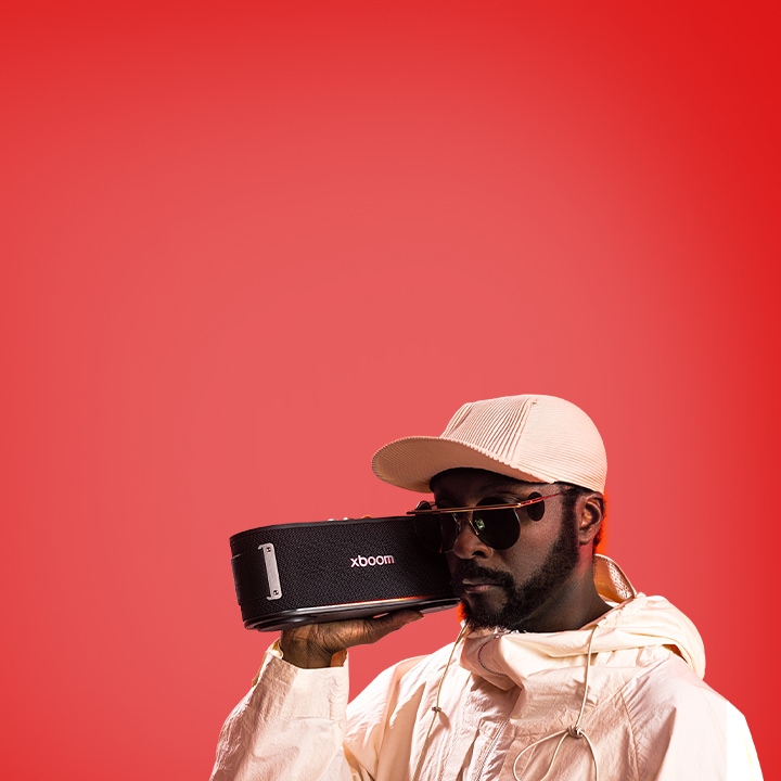 will.i.am beyaz kıyafet ve güneş gözlüğüyle, xboom Bounce’u yüzünün hemen yanında tutuyor.