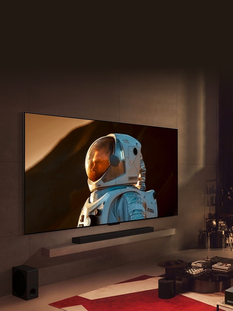 一台超大型 LG TV 安裝在溫馨客廳的墻上，顯示宇航員的特寫畫面佔據了墻面的大部分區域。一名男士和寵物狗坐在電視前方的沙發上看電視，窗外呈現夜晚的城市風光。