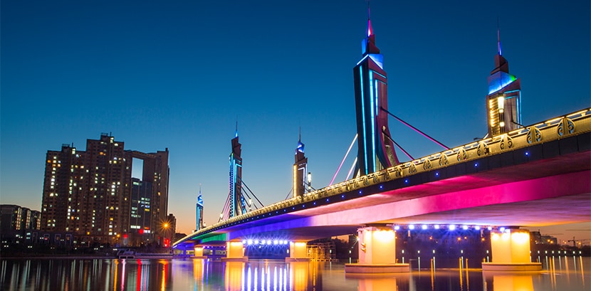 燈火通明的現代橋樑和摩天大樓的夜景，水中倒映著鮮豔的燈光，展現高對比度和色彩細節。	