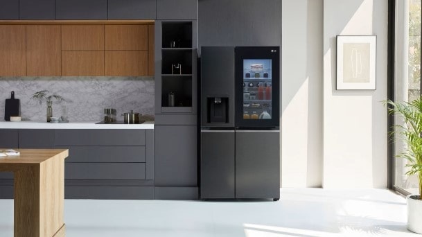 LG InstaView American-style fridge freezer with illuminated door panel in a modern kitchen with dark cabinets, marble backsplash, and natural light from a large window.