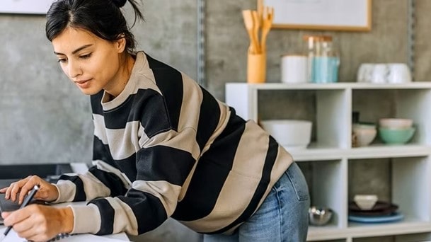 Woman in striped shirt leaning over a table while writing, in a modern kitchen setting with open shelving and ceramics in the background.