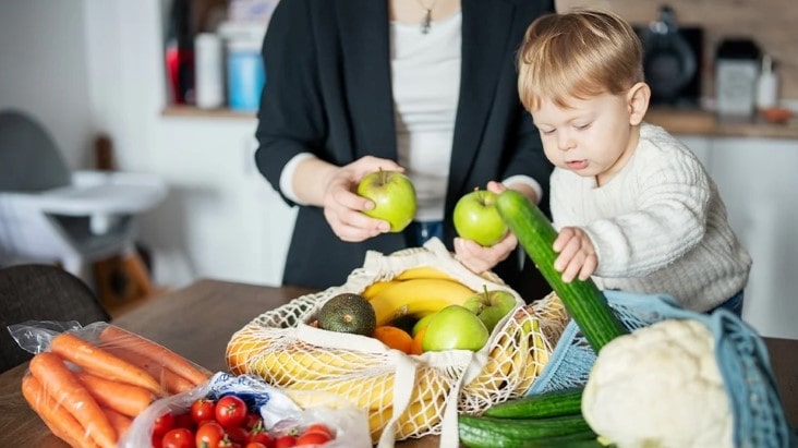 Toddler and adult sorting fresh fruits and vegetables on a kitchen table, symbolising grocery needs when choosing the right fridge freezer size.
