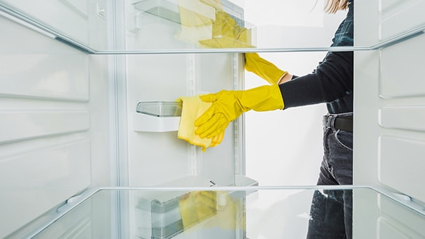 A person wearing yellow gloves is cleaning the inside of a refrigerator with a yellow cloth, seen from an interior view