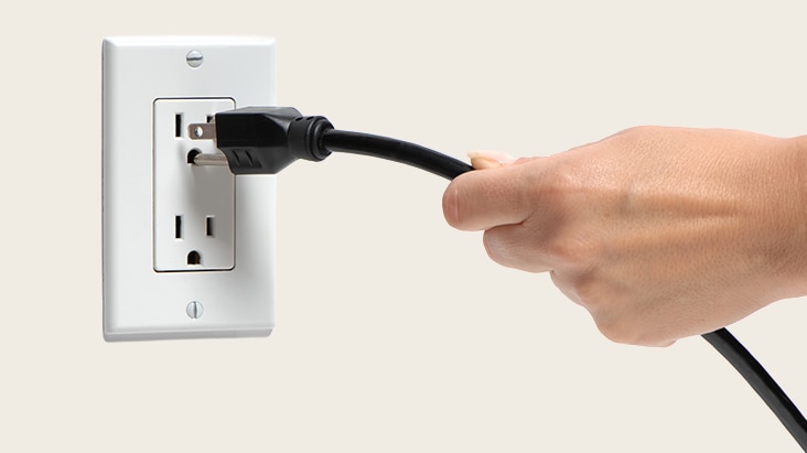 A hand unplugging a black power cord from a white electrical outlet on the wall.