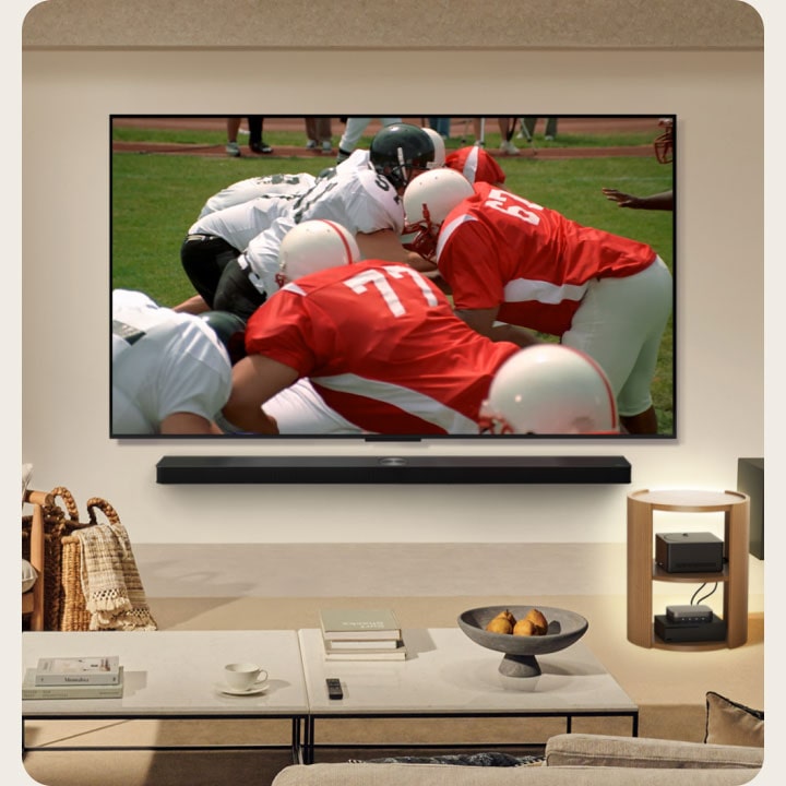 Living room with an LG True Wireless TV on the wall. On the screen is an American football match. The TV setup also includes a wall-mounted LG Soundbar. And almost hidden from sight, the other peripherals are connected to a Zero Connect Box that is on a side table. The whole living room is neat, tidy, and has no wire-clutter. 