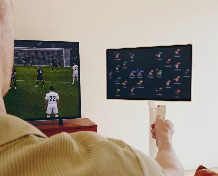 The back of a man holding a remote control towards a TV showing a football match and an LG Smart Monitor Swing smart screen displaying a match analysis.
