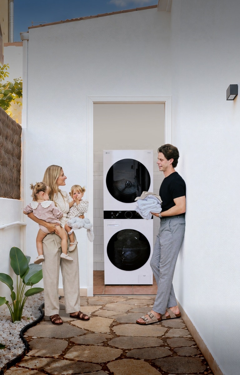 A family with toddler twins standing in a white-walled outdoor hallway next to a stacked LG washing machine and tumble dryer unit.