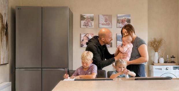 A family gathers in a kitchen near a stainless steel LG refrigerator—a boy writes at the dining table, a young girl sits beside him with a small bottle, and parents stand together holding a baby.