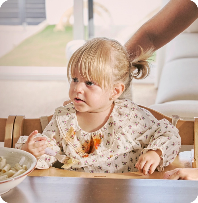 A young toddler sitting in a wooden high chair, holding a spoon with food stains on their floral outfit.