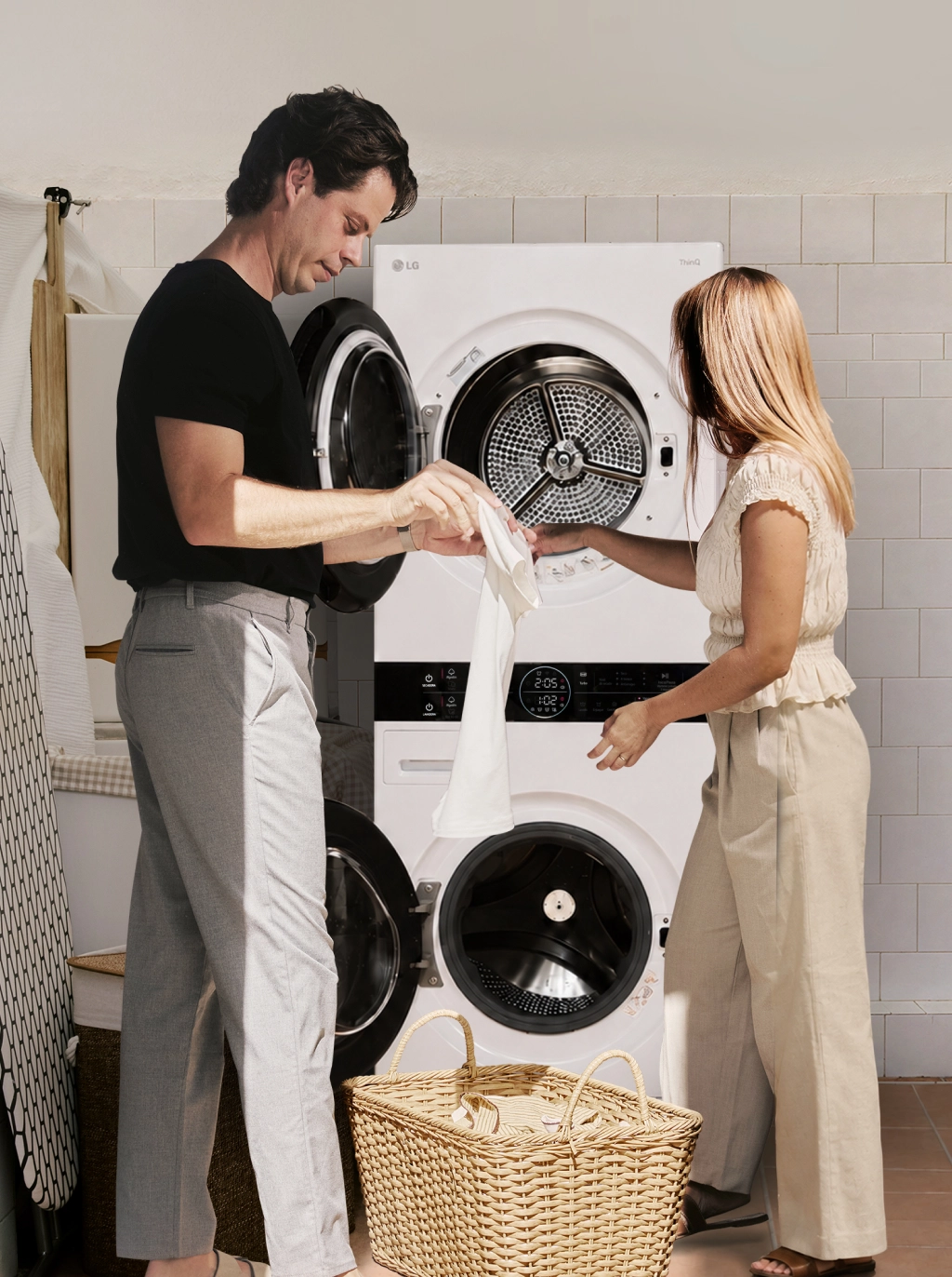 A man and woman transferring white laundry between a stacked washing machine and tumble dryer in a utility room.