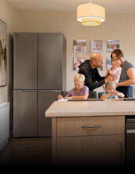 A family gathers in a kitchen near a stainless steel LG refrigerator—a boy writes at the dining table, a young girl sits beside him with a small bottle, and parents stand together holding a baby.