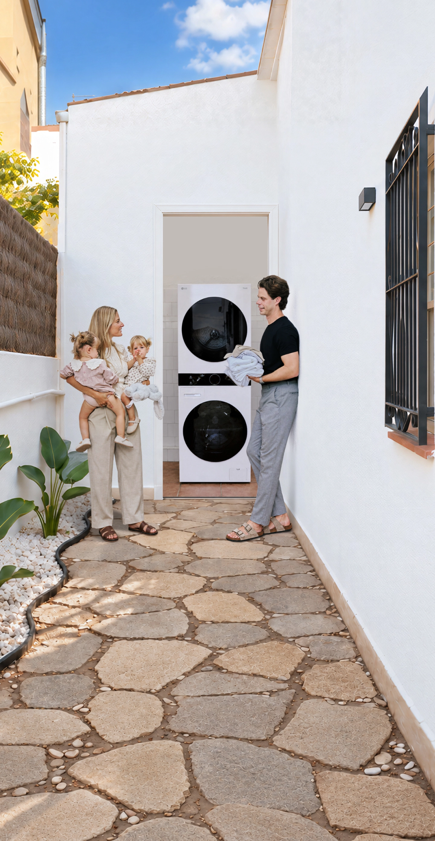 A family with toddler twins standing in a white-walled outdoor hallway next to a stacked LG washing machine and tumble dryer unit.