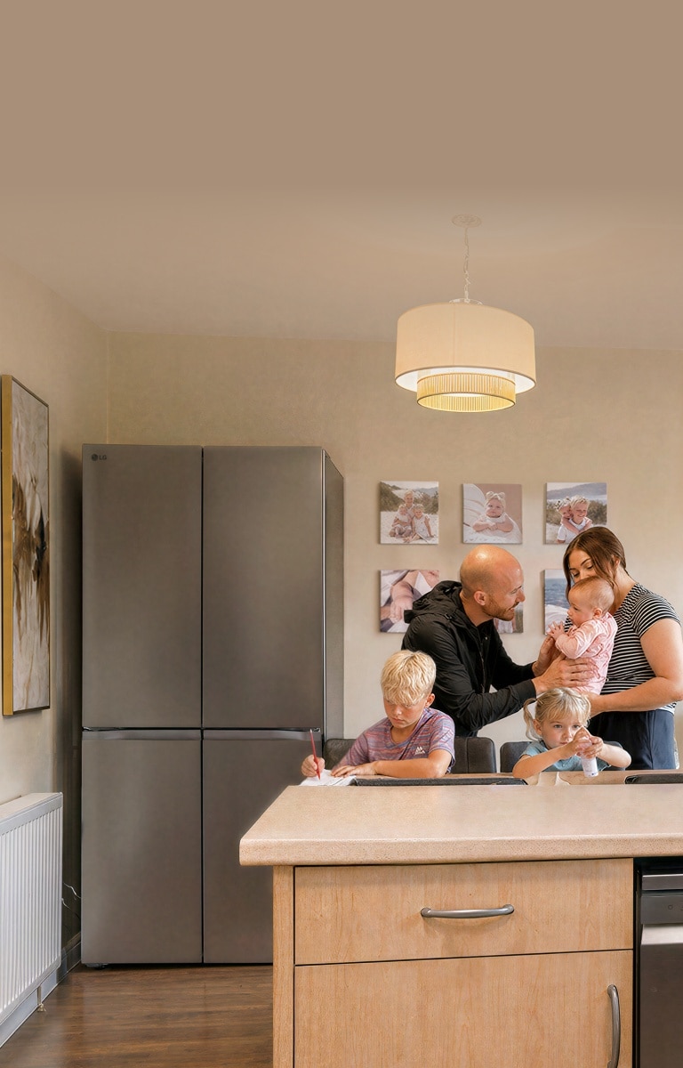 A family gathers in a kitchen near a stainless steel LG refrigerator—a boy writes at the dining table, a young girl sits beside him with a small bottle, and parents stand together holding a baby.