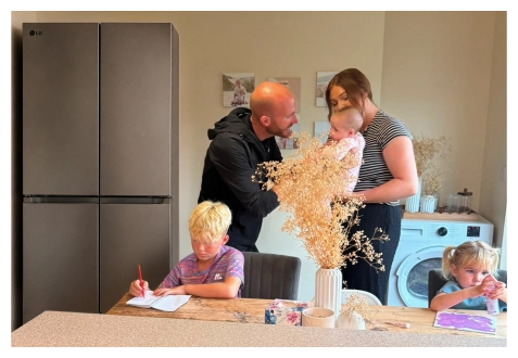 A family gathers in the kitchen near a stainless steel refrigerator—a boy draws at the table, a girl paints at the counter, and parents stand together holding a baby.