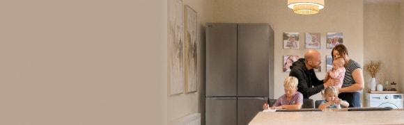 A family gathers in a kitchen near a stainless steel LG refrigerator—a boy writes at the dining table, a young girl sits beside him with a small bottle, and parents stand together holding a baby.
