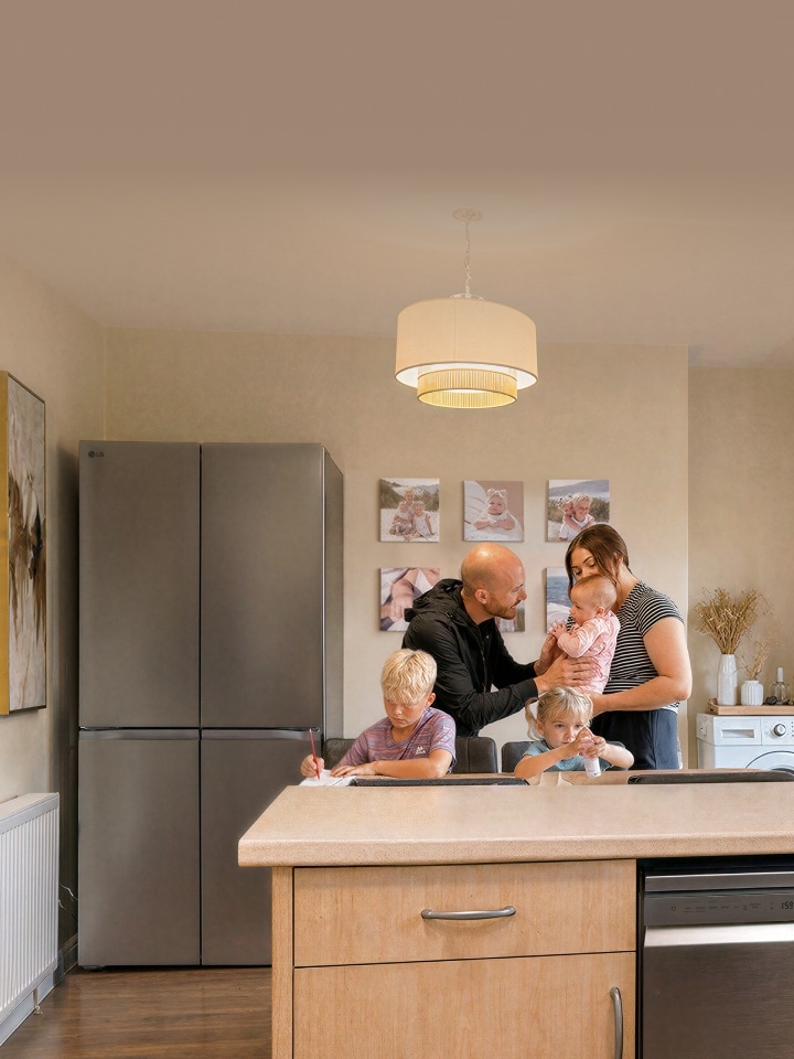 A family gathers in a kitchen near a stainless steel LG refrigerator—a boy writes at the dining table, a young girl sits beside him with a small bottle, and parents stand together holding a baby.