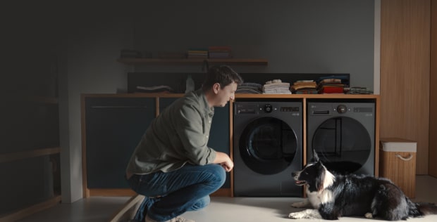 In a home laundry room, a man and a dog are facing each other, with two matte black LG washing machines visible directly behind them.
