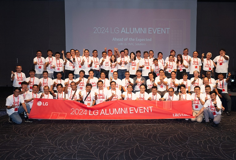 Group of people posing for a photo  at the 2024 LG Alumni Event, holding a banner that reads '2024 LG Alumni Event' in front of a presentation screen.	