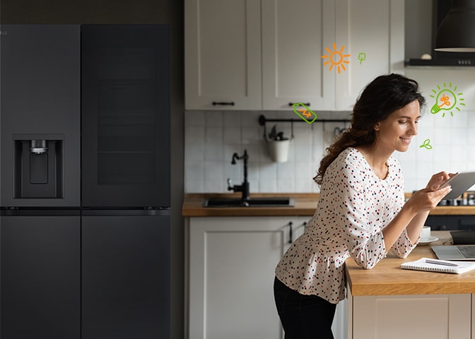 In front of the refrigerator, the woman is smiling while looking at the tab, and there are light bulbs and nature icons around the woman that represent energy efficiency.