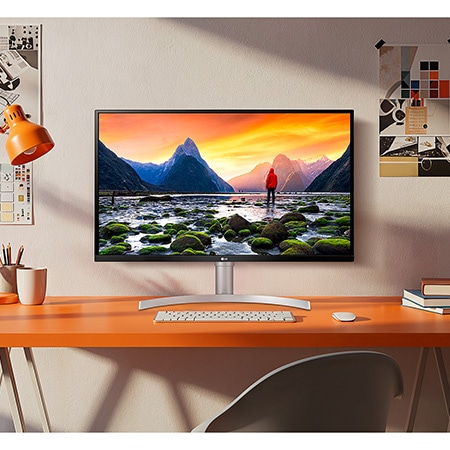 The LG Monitor 32UN650K in a white/silver finish, featuring UHD 4K resolution, displays a brilliant landscape image in a stylish, creative setting on an orange desk.