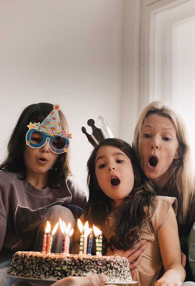 Image of two adult women and a young girl wearing a birthday hat on their head and blowing out the candles on the cake.