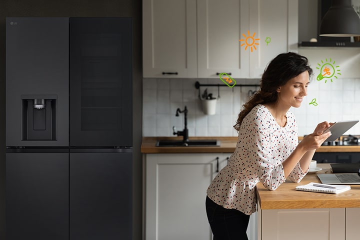 In front of the refrigerator, the woman is smiling while looking at the tab, and there are light bulbs and nature icons around the woman that represent energy efficiency.