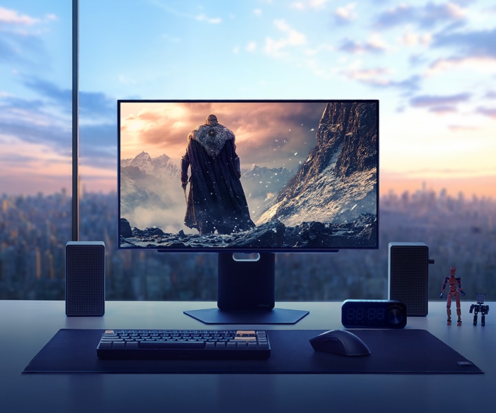 A high-end gaming monitor displays a dramatic fantasy game scene featuring a cloaked warrior standing before a snowy mountain range. The monitor is set on a sleek desk in a high-rise apartment with a city skyline at sunset in the background. Surrounding the monitor are speakers, a keyboard, headset, clock, and small action figures, showcasing an immersive and stylish gaming setup.