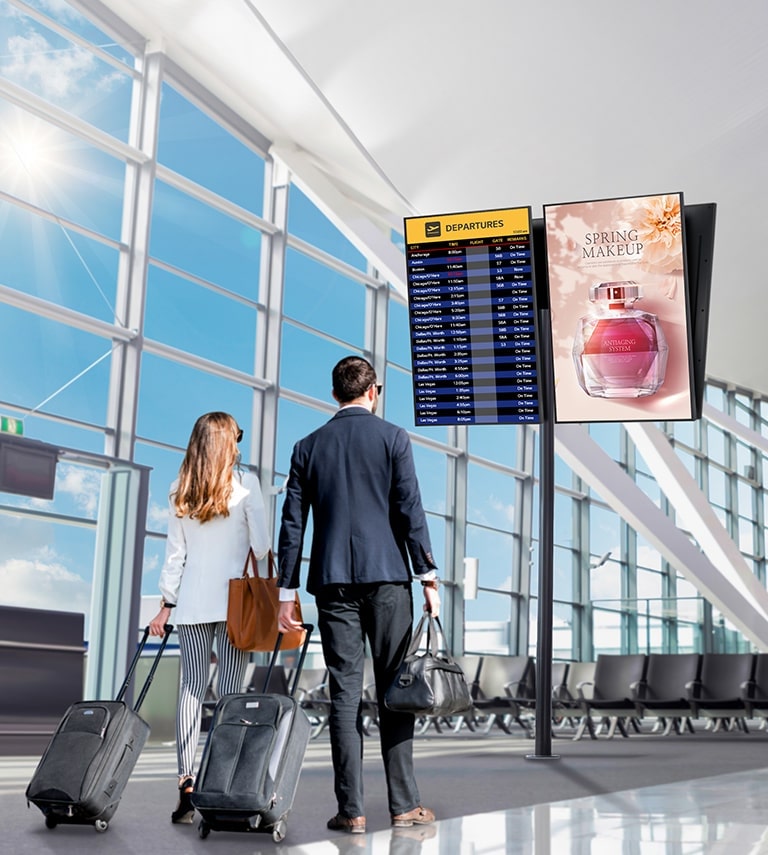 A man and a woman are checking the departure time of the flight through the signage installed at the airport. And the screen with anti-glare coating has small reflection of the light from the sun.