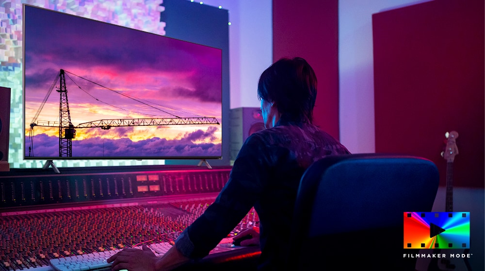 A movie director is looking at a big TV monitor, editing something. The TV screen shows a tower crane in purple sky. FILMMAKER Mode logo is placed on bottom right corner.