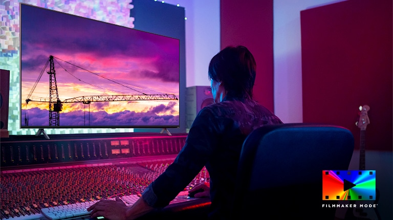 A movie director is looking at a big TV monitor, editing something. The TV screen shows a tower crane in purple sky. FILMMAKER Mode logo is placed on bottom right corner. 