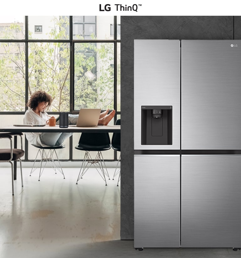 Modern kitchen with LG ThinQ fridge, person working on laptop at table by large windows with greenery outside.