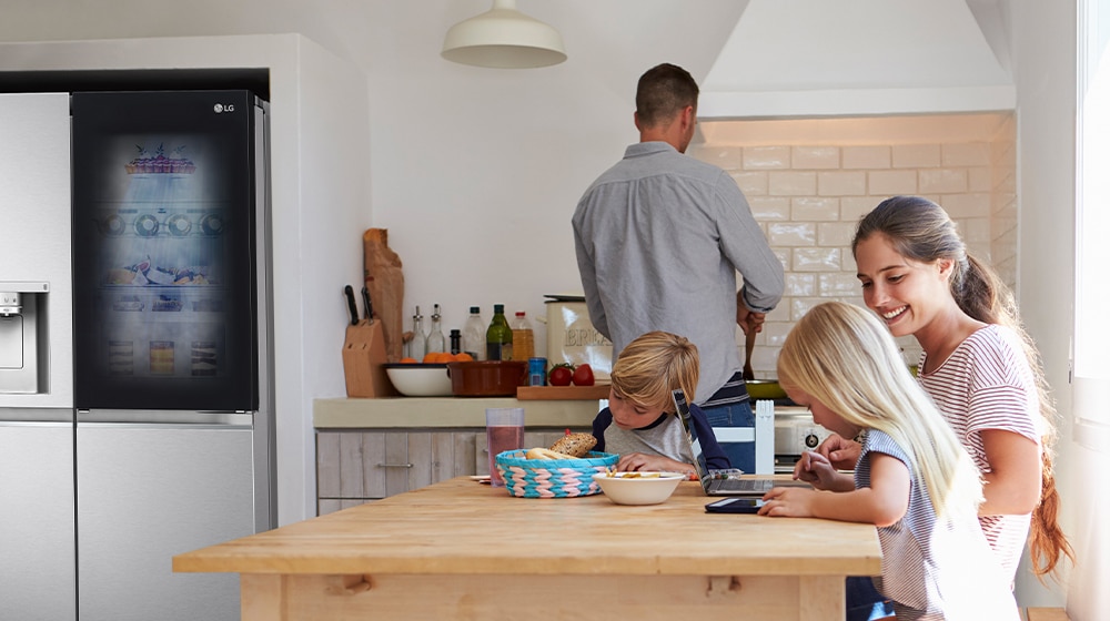 A family in a kitchen with an LG refrigerator; a man cooks while two children use laptops at a wooden table.