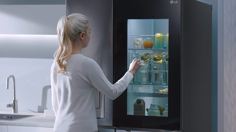 A video shows a woman approach her InstaView refrigerator and knock twice. The interior lights up and she can see the contents of her fridge without opening the door. The view zooms in to focus on the drinks in the door and then zooms out to see the woman from behind as she opens the door and grabs a drink.