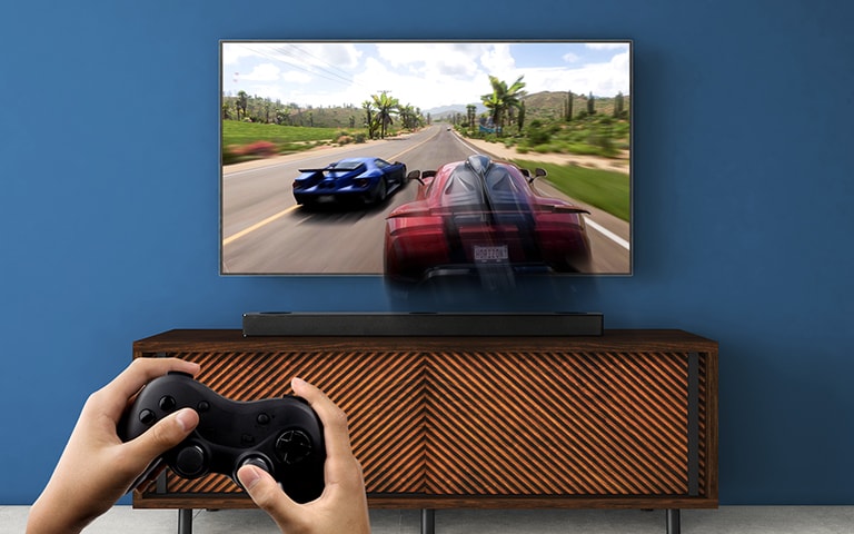 LG TV is on the wall, showing a racing game. LG Sound Bar is place on the brown shelf, right below LG TV. A man is holding a joy stick.