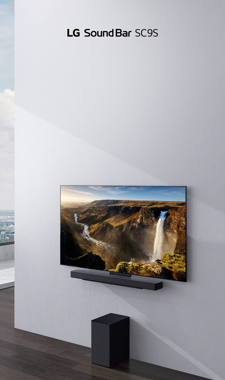 TV and LG Soundbar  SC9S are hung on a white wall. Below a black wireless subwoofer is placed on the floor. Beyond the window on the left a city view with the blue sky can be found.