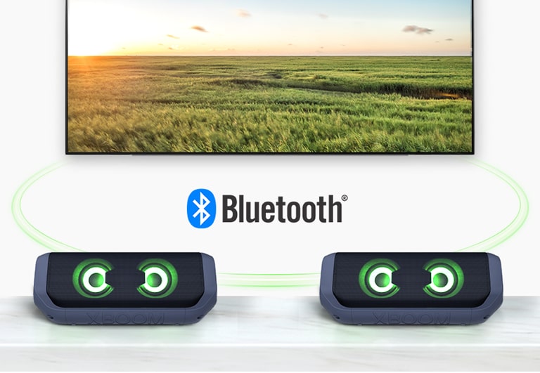 On a table, two LG XBOOM Go with green lighting are in front of a TV showing a field.