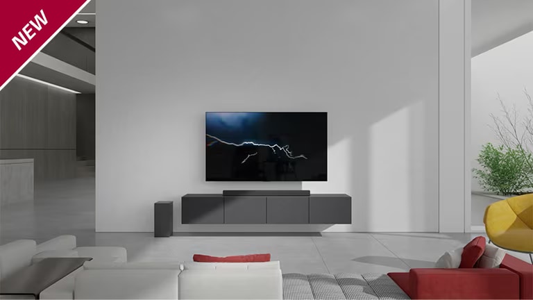 The sound bar is placed on gray cabinet with a TV hung on the wall in the living room. A wireless subwoofer is placed on the floor on the left side and the sunlight comes in from the right side of the picture. A white and red colored long sofa is placed facing the TV and sound bar. NEW mark is shown in the top left corner.