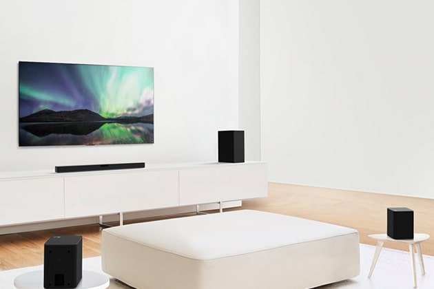 TV and sound bar in white living room with white sofa in the center. Speakers sit on both ends of the sofa.